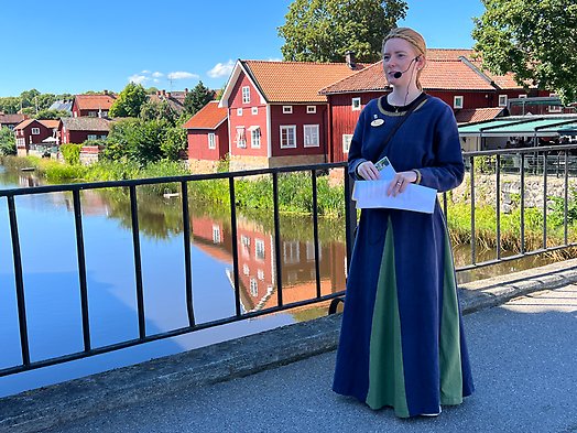 Guide i medeltidsklänning på Kapellbron i Arboga i sommargrönska.