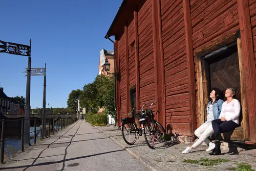 Ladbron vid Arbogaån, två kvinnor sitter vid rött hus i vårsolen.